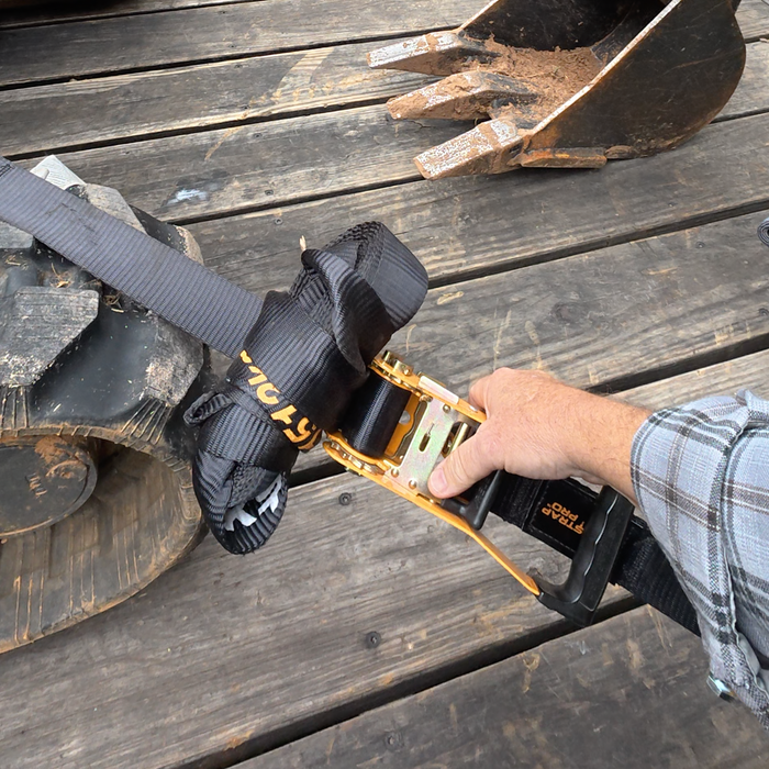 Hand operating a gold TorkStrap PRO ratchet beside an excavator track on a wooden trailer.